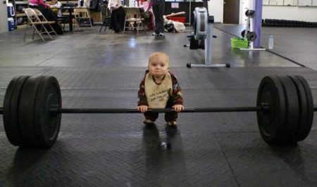 Photo showing a baby with hands on a huge barbell in a gym