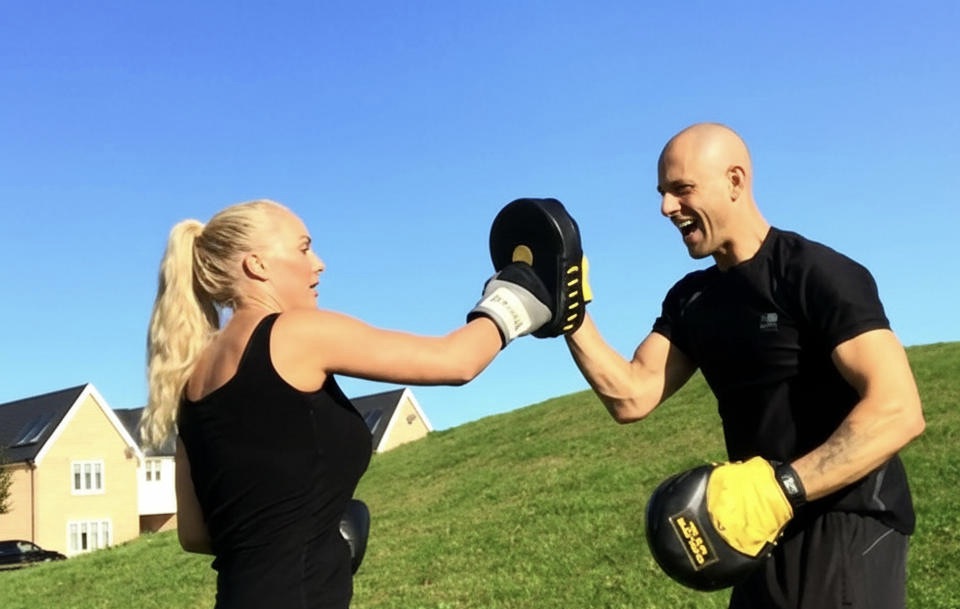 Andy holding focus pads for a female client while she hits them, outside on a grassy verge with a blue sky